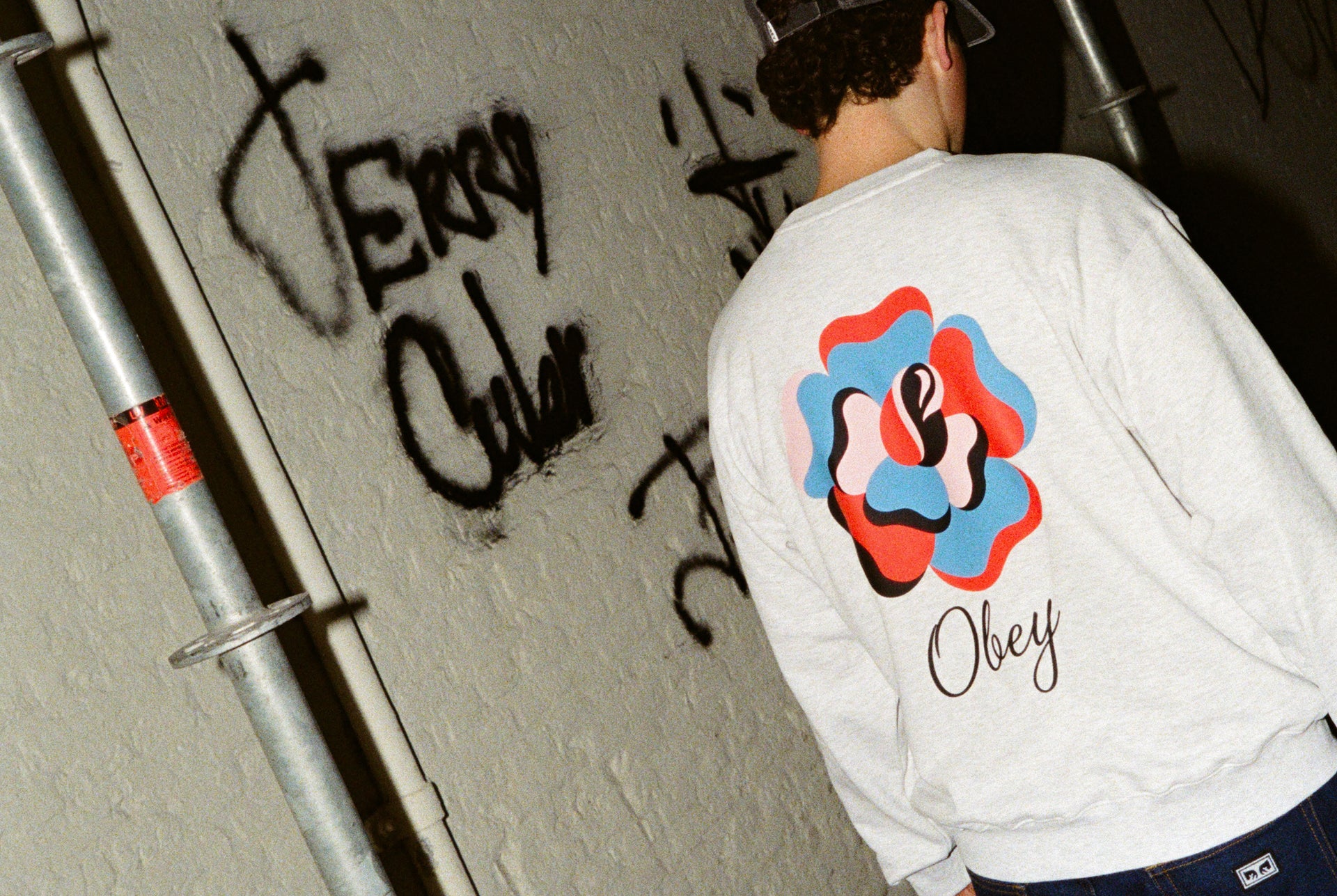 Person wearing a white sweatshirt with a colorful logo and 'Obey' text, standing against a wall with graffiti.