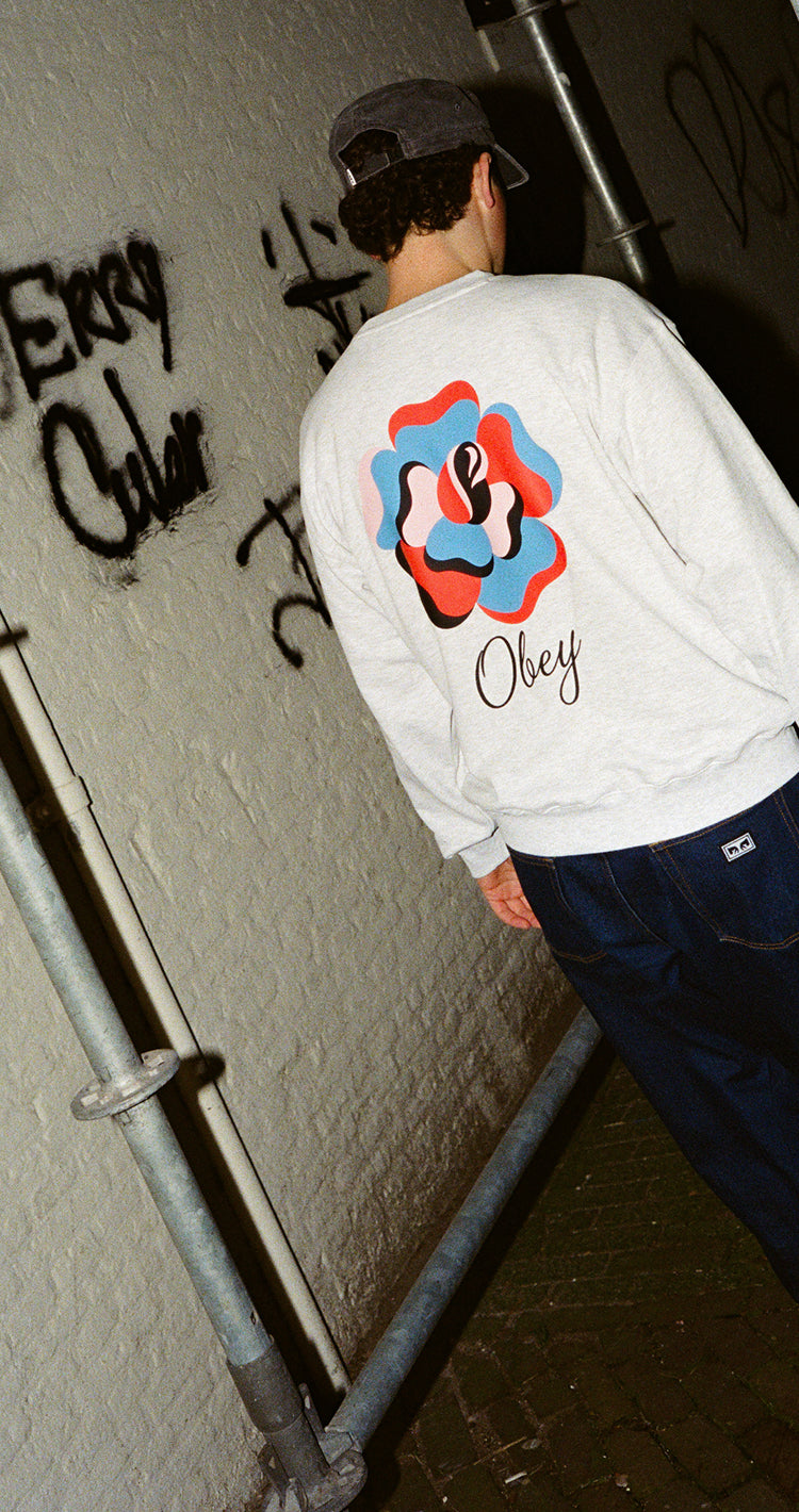 Person wearing a white sweatshirt with a colorful design and text, standing against a wall with graffiti.
